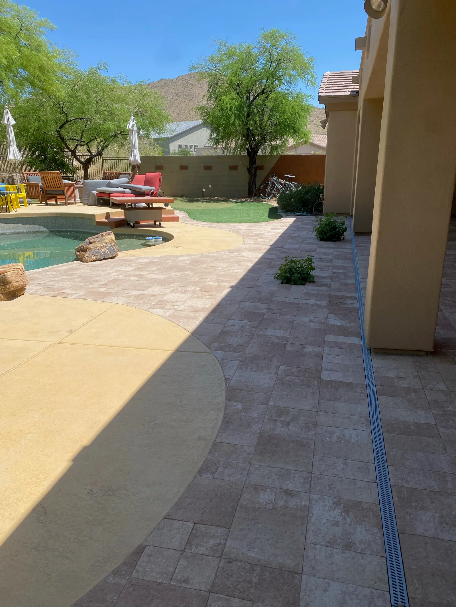 Concrete patio with dark pavers, drainage channel, curved edges, and pool area with landscaping